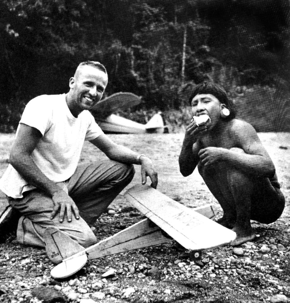 Nate Saint with a Waorani man and a model airplane on a riverbank — a moment of friendship before Palm Beach